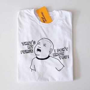 May include: White t-shirt with a black and white illustration of a cartoon character with the text "That's My Purse I Don't Know You!"