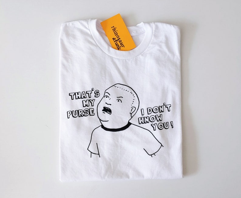 May include: White t-shirt with a black and white illustration of a cartoon character with the text "That's My Purse I Don't Know You!"