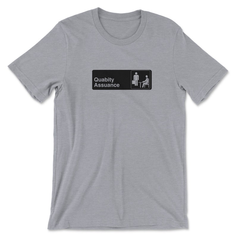 May include: A heather grey t-shirt featuring a black and white graphic of a sign that reads "Quality Assurance" with two stick figures, one standing and one sitting at a desk.