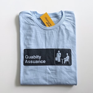 May include: Light blue t-shirt featuring a black and white graphic of a sign that reads "Quality Assurance" with two stick figures. The stick figure on the left is holding a briefcase and the stick figure on the right is sitting at a desk.