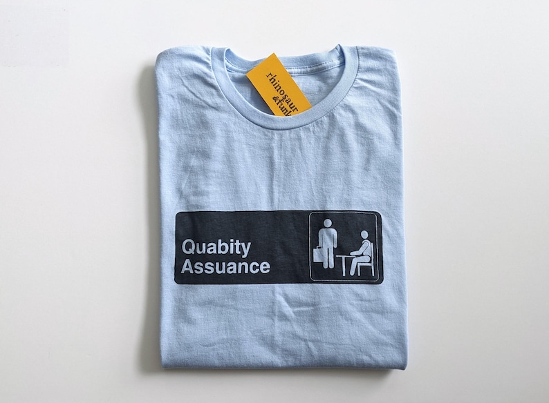 May include: Light blue t-shirt featuring a black and white graphic of a sign that reads "Quality Assurance" with two stick figures. The stick figure on the left is holding a briefcase and the stick figure on the right is sitting at a desk.