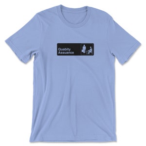 May include: Light blue t-shirt featuring a black and white graphic of a sign that reads "Quality Assurance" with a stick figure holding a briefcase and another stick figure sitting at a desk.