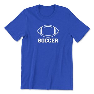 May include: A royal blue t-shirt featuring a white graphic of a football shape with the word "SOCCER" printed below it in a bold, sans-serif font. The shirt is made of a soft material.