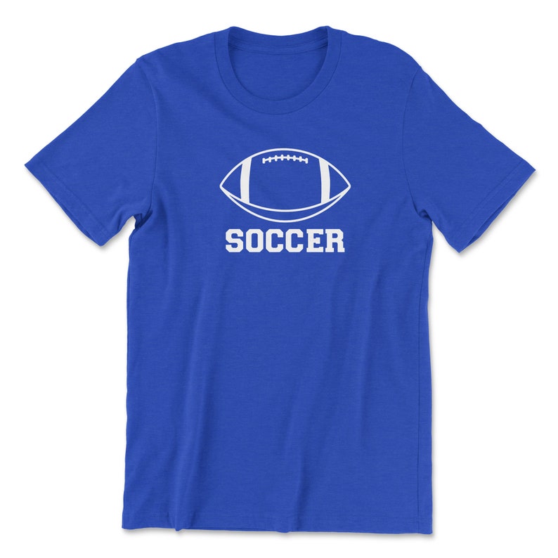 May include: A royal blue t-shirt featuring a white graphic of a football shape with the word "SOCCER" printed below it in a bold, sans-serif font. The shirt is made of a soft material.