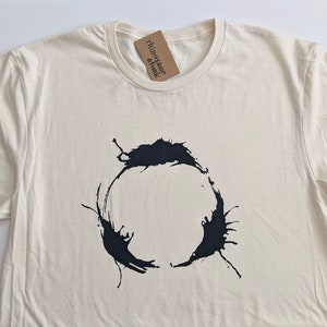 May include: A cream colored t-shirt with a black ink splatter design that resembles a circle. The design is centered on the shirt. The shirt has a crew neck and short sleeves. A small brown tag with the text "rhinosaure & funk" is attached to the shirt.
