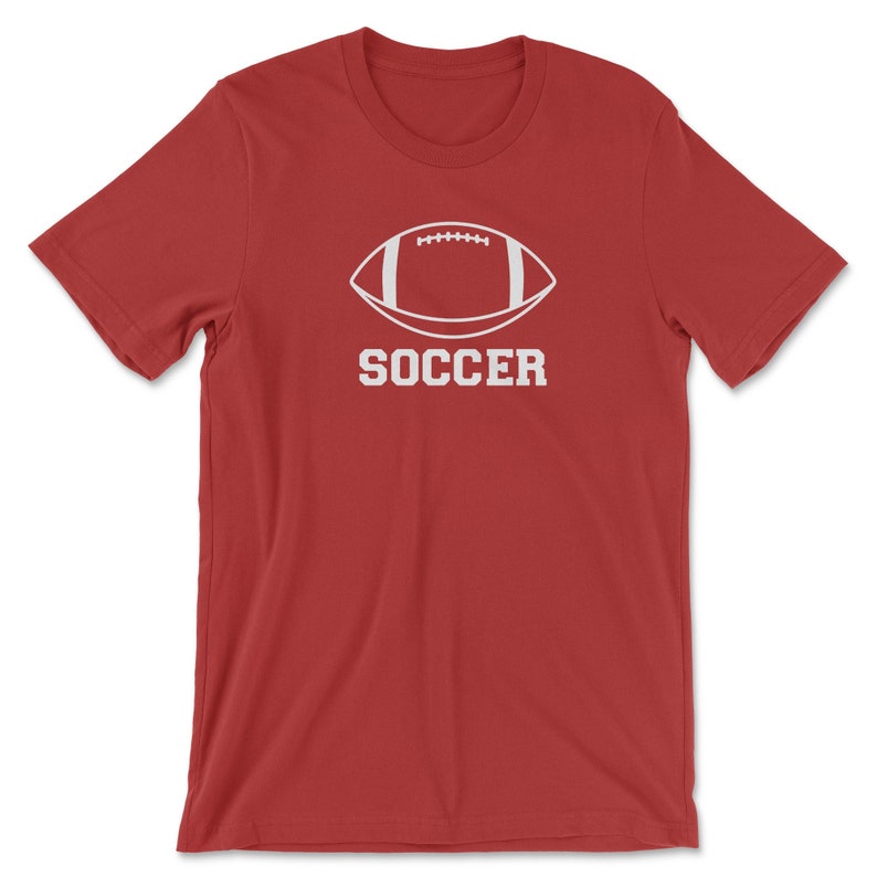 May include: A red t-shirt featuring a white graphic of a football shape with the word "SOCCER" printed below it in a bold, sans-serif font. The shirt is made of a soft material and has short sleeves.