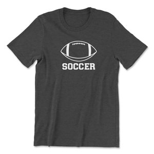 May include: Dark gray t-shirt featuring a white graphic of a football shape with the word "SOCCER" in bold, capital letters underneath. The shirt has short sleeves and a crew neck.