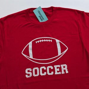 May include: Red t-shirt with a white football graphic and the word "SOCCER" printed below. The shirt has a tag that reads "rhinosaur & funk".