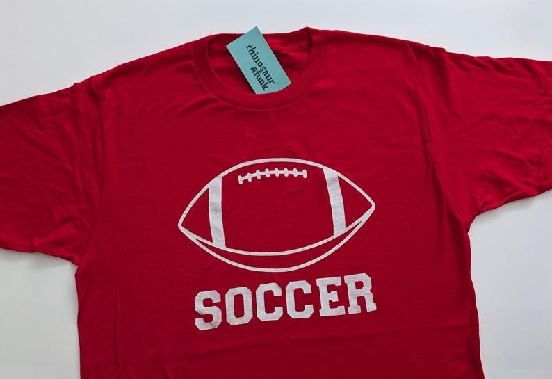 May include: Red t-shirt with a white football graphic and the word "SOCCER" printed below. The shirt has a tag that reads "rhinosaur & funk".