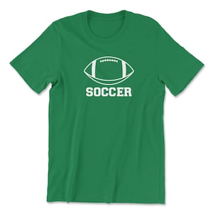 May include: A green t-shirt featuring a white graphic of a football with the word "SOCCER" printed below it in a bold, sans-serif font. The shirt is made of a soft material and has short sleeves and a crew neck.