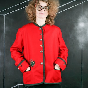 May include: A red jacket with black trim and a green heart-shaped design on the front. The jacket has a button-up closure and pockets on the sides.