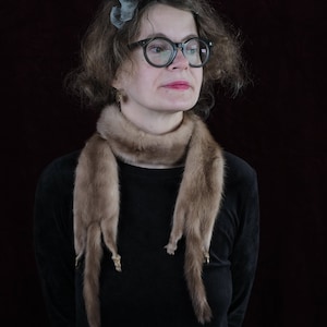 May include: A woman with curly brown hair is wearing a black turtleneck sweater and a brown fur scarf. She is wearing glasses and has a small, silver earring in her left ear.