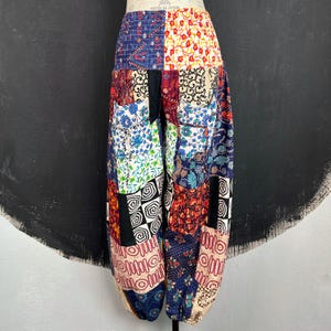 May include: Patchwork trousers featuring a mix of patterns and colours, such as floral, geometric, and paisley. The trousers have a relaxed fit and a gathered waistband. The colour scheme includes blue, red, white, and black.