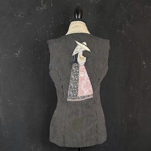 May include: A black and white striped vest with a decorative applique of a woman in a hat and dress. The applique features floral and patterned fabrics in shades of pink, blue, and white. The vest is displayed on a mannequin.