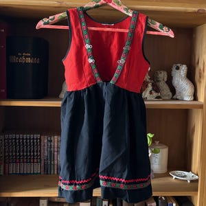Könnte beinhalten: Ein ärmelloses Kleid mit rotem Oberteil und schwarzem Rock. Das Oberteil hat einen tiefen V-Ausschnitt, der mit einem dekorativen grün-roten Musterband und zwei silberfarbenen Metallverschlüssen verziert ist. Der schwarze Rocksaum hat eine Borte.