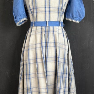 May include: A white and blue plaid dress with puffed blue sleeves and a blue belt. The dress is on a mannequin against a black background.