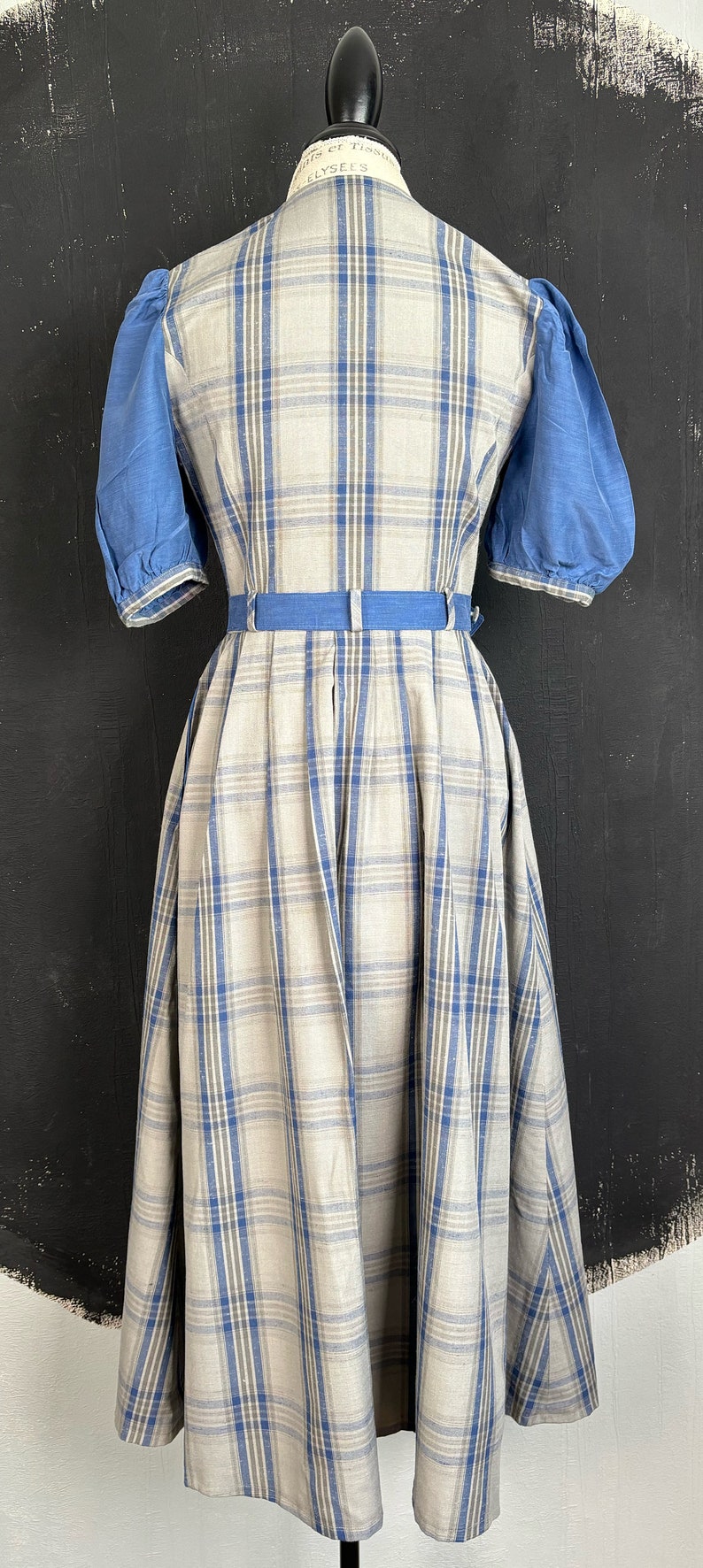 May include: A white and blue plaid dress with puffed blue sleeves and a blue belt. The dress is on a mannequin against a black background.