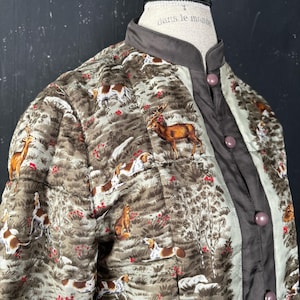May include: A quilted jacket featuring a brown and green woodland print with deer and hunting dogs. The garment has a solid dark brown mandarin collar, a front button placket with matching round buttons, and a light-colored interior lining.