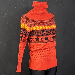May include: Orange turtleneck sweater with a repeating pattern of black diamonds, yellow flowers, and apple logos. The sleeves and lower portion of the sweater have a repeating pattern of letters. The sweater is made of a soft, knitted material.