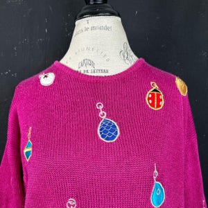 Puede incluir: Un suéter de punto magenta con adornos bordados. Los adornos son una mariquita, una concha y una estrella.