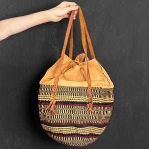Puede incluir: Un bolso tote tejido con una parte superior de cuero color canela con cordón y correas de cuero. La parte inferior de la bolsa presenta un patrón geométrico negro, rojo y canela. La bolsa está sostenida por una mano sobre un fondo oscuro.