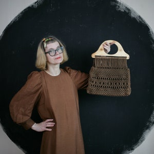 May include: A brown macrame handbag featuring a complex knotted pattern and a light-colored, semi-circular wooden handle. The bag is constructed from dark brown cord, showcasing a detailed woven texture throughout the body of the accessory.