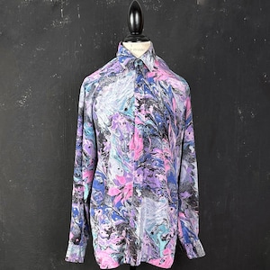 Puede incluir: Una camisa de manga larga con botones y un estampado abstracto colorido en tonos de rosa, azul, morado y negro. La camisa tiene un cuello clásico y un cierre de botones.