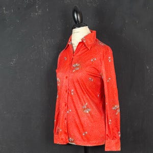 May include: A long-sleeved button-down shirt in a vibrant red hue, featuring a delicate white polka dot pattern throughout and small, scattered floral motifs. The shirt includes a classic pointed collar and buttoned cuffs.