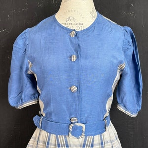 May include: A blue and white linen dress with a plaid skirt and a solid blue bodice. The dress has a button-up front and a belt with a buckle.