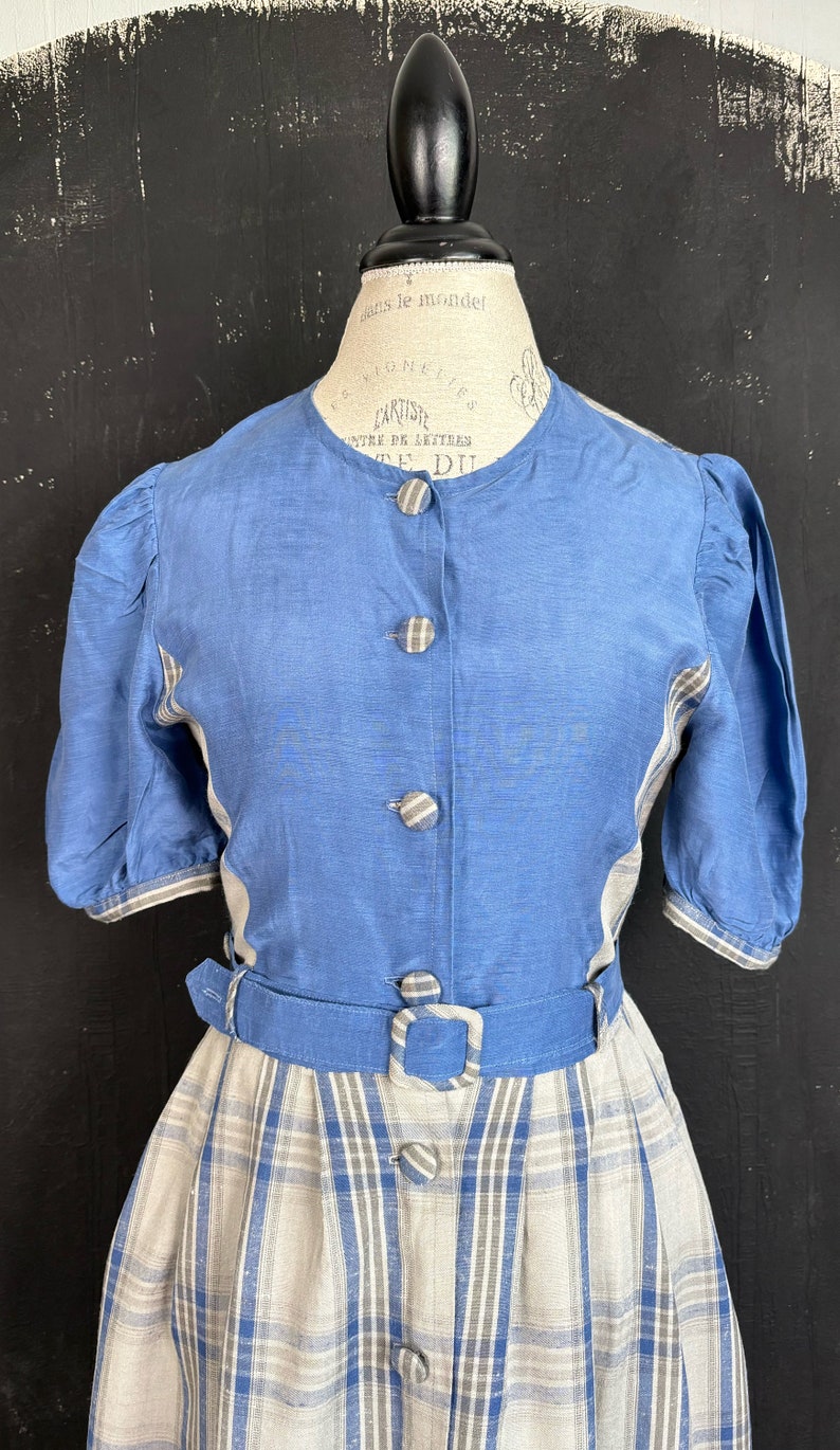 May include: A blue and white linen dress with a plaid skirt and a solid blue bodice. The dress has a button-up front and a belt with a buckle.