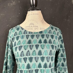 Puede incluir: Una blusa de manga larga de color verde azulado apagado, adornada con un patrón repetido de formas de hojas verdes y turquesas estilizadas. La blusa se exhibe en un maniquí.