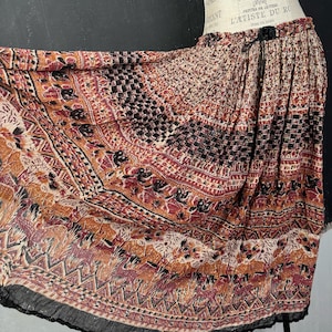 May include: A flowing skirt with an intricate pattern in shades of brown, black, and red. The fabric is gathered at the waist and falls in soft folds. The design features geometric shapes and stylized figures, with a black lace trim at the hem.