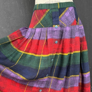 May include: A pleated skirt featuring a multi-colored plaid pattern in red, green, navy blue, purple, and yellow. The skirt has an elasticized waistband and a vertical front placket secured with five small, round, teal-colored buttons.