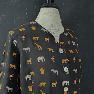 Könnte beinhalten: Schwarzes Hemd mit einem sich wiederholenden Muster von Safari-Tieren in Gold und Weiß. Die Tiere umfassen Elefanten, Giraffen, Zebras, Löwen und Flamingos. Das Hemd hat drei große, runde, irisierende Knöpfe.