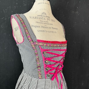 Op de afbeelding: Een mouwloze jurk met een zwart-wit gingham patroon, geaccentueerd met felroze biezen en een decoratieve bloemenlintrand. Het lijfje heeft een vetersluiting aan de voorzijde met hartvormige metalen oogjes en een roze lint.