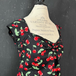 Op de afbeelding: Zwarte mouwloze top met een kersenprint. De top heeft een vierkante halslijn en een getailleerde lijfje. De stof is zwart met een patroon van rode kersen en groene bladeren. De top wordt op een mannequin getoond.