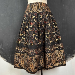 Op de afbeelding: Zwarte midi-rok met een paisley patroon in goud, bruin en rood. De rok heeft een brede tailleband en een geplooid ontwerp, waardoor een volle, vloeiende silhouet ontstaat. De zoom is versierd met een decoratieve rand. Ongeveer 76 cm lang.