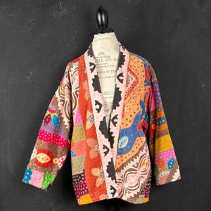 Può includere: Una giacca patchwork con un vivace mix di colori e motivi. La giacca presenta una varietà di tessuti e disegni, tra cui forme floreali e geometriche. La giacca è esposta su un manichino.