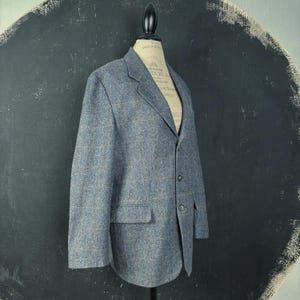 Puede incluir: Un blazer de tweed azul con un sutil estampado de cuadros, exhibido en un maniquí. La chaqueta presenta solapas con muescas, dos bolsillos delanteros y tres botones. La tela tiene una apariencia texturizada.
