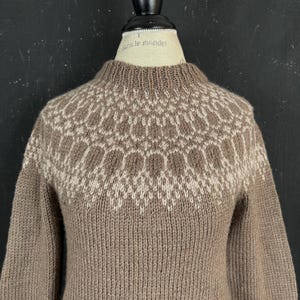 May include: A long-sleeved, brown knit jumper featuring a circular yoke with a cream-coloured geometric pattern. The garment has a ribbed crew neckline and is displayed on a mannequin with the text "dans le monde" printed on the neck area.
