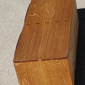 May include: A handcrafted wooden box with a natural wood grain finish. The box has a unique, angled design and visible dovetail joints. The warm brown tones of the wood are highlighted by the light. The box is likely used for storage or display.