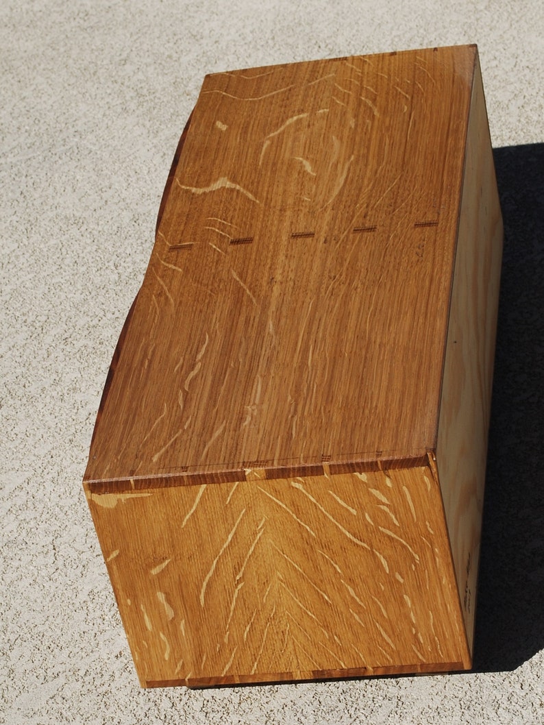 May include: A handcrafted wooden box with a natural wood grain finish. The box has a unique, angled design and visible dovetail joints. The warm brown tones of the wood are highlighted by the light. The box is likely used for storage or display.