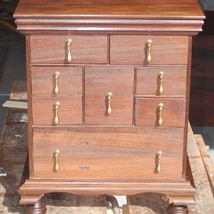 May include: A small, wooden chest of drawers with a dark brown finish. It features eight drawers with brass-colored teardrop pulls. The chest rests on four rounded feet and has a decorative top.