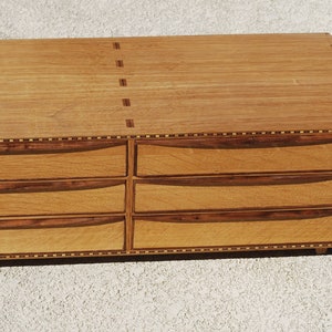 May include: A handcrafted wooden dresser with six drawers. The dresser is made of light-colored wood with darker wood accents on the drawer fronts and edges. A blue baseball cap with white lettering is in the lower right corner.