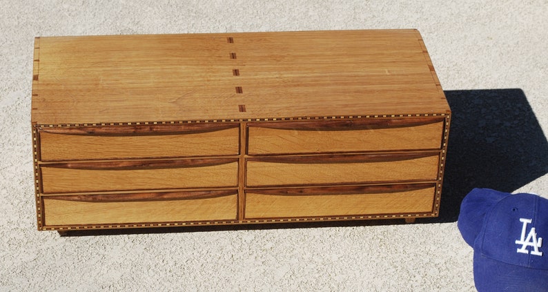 May include: A handcrafted wooden dresser with six drawers. The dresser is made of light-colored wood with darker wood accents on the drawer fronts and edges. A blue baseball cap with white lettering is in the lower right corner.
