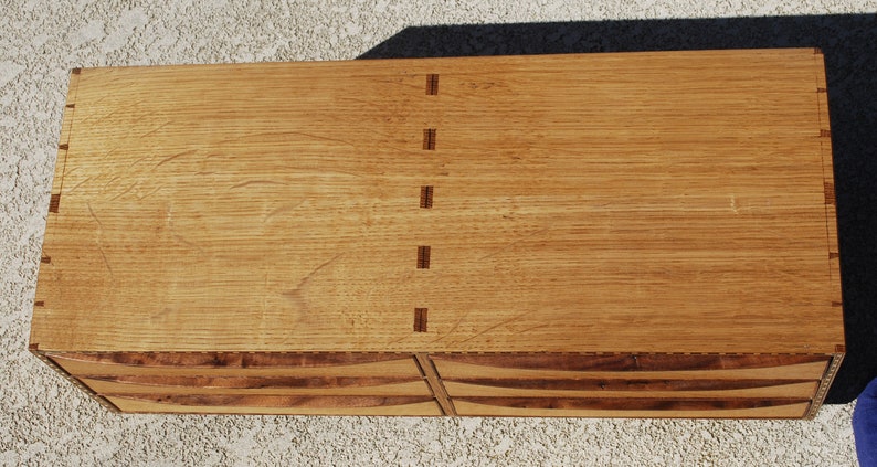 May include: A handcrafted wooden chest with a light brown finish. The chest features dovetail joints and a series of curved drawers. The wood grain is visible, adding to the natural aesthetic. The chest is rectangular in shape.