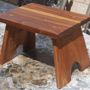 May include: A small, handcrafted wooden stool with a rectangular top and angled legs. The stool is made of dark brown wood with visible grain and features small, contrasting wood accents. The design is simple and sturdy.