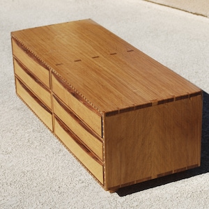 May include: A handcrafted wooden dresser with six drawers. The dresser is made of light brown wood with darker wood accents around the drawer fronts and edges. The drawers have curved fronts and are arranged in two columns of three.