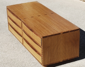 May include: A handcrafted wooden dresser with six drawers. The dresser is made of light brown wood with darker wood accents around the drawer fronts and edges. The drawers have curved fronts and are arranged in two columns of three.