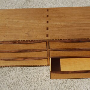May include: A wooden chest of drawers with six drawers, one partially open. The chest is made of light brown wood with darker brown accents and decorative inlays. The drawers have curved handles and the chest is rectangular.